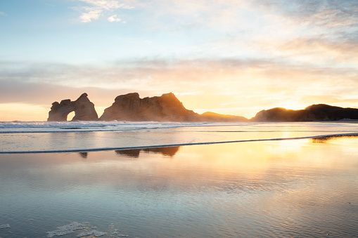 Sunrise on Wharariki Beach, New Zealand, looking towards the Archway Islands just off the coast. The pastel colored sky is reflected in the sand and gentle waves. \nThis beach is a popular tourist destination and is known for it's beautiful reflections of the islands.