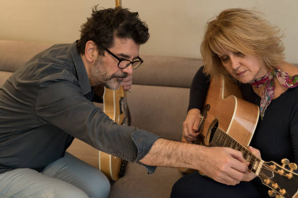 istock A middle-aged couple sits on a sofa next to each other in a cozy environment. They are both holding guitars, having a good time and laughing. The men is teaching the woman how to play. 1287782201
