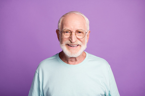 Portrait of smiling optimistic beard pensioner man wear light blue t-shirt eyeglasses isolated on purple color background.