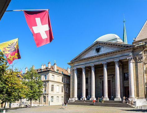 Geneva, Switzerland - September 5, 2020: Neoclassical facade of St. Pierre cathedral in the old town, home church of John Calvin, with a swiss flag and a flag of Geneva hanging against blue sky.