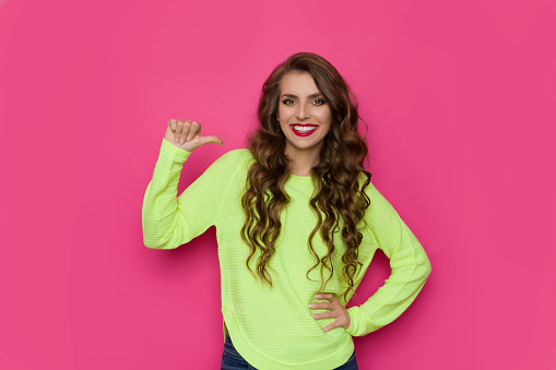 Smiling young woman in neon green sweater is pointing with thumb at herself. Waist up studio shot on pink background.