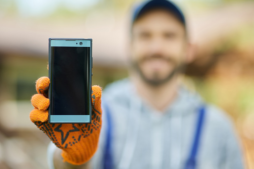 Close up of smartphone with blank screen. Young male builder in uniform using mobile phone, standing outdoors on a warm sunny day. Technology concept. Selective focus