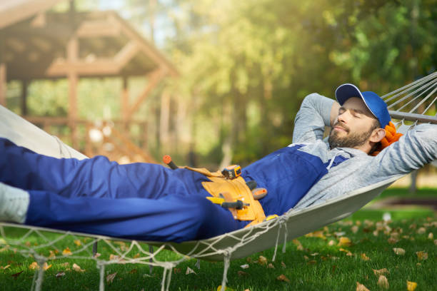 Relaxed young male builder wearing blue overalls and cap taking a break, lying in a hammock outdoors with eyes closed on a sunny day
