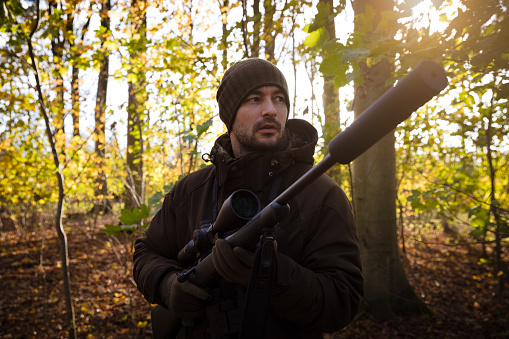 Male hunter with rifle and binocular stalking roe deer in beautiful autumn forest in Denmark. During covid-19 related lockdown from work, more people spend time with their hobbies and pursuing outdoor activities and a healthy lifestyle.