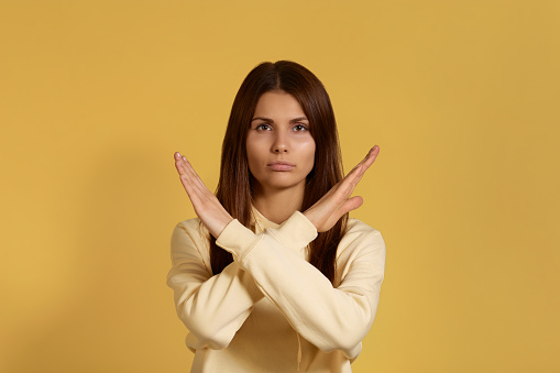 Cant go further. Serious caucasian woman in yellow hoodie keeps arms crossed, makes stop or ban gesture, indicates that the passage is closed. Isolated on a yellow background