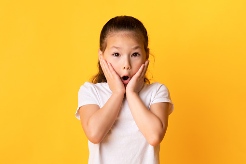 Wow. Portrait of surprised and shocked asian girl touching cheeks with open mouth in amazement isolated over yellow studio background. Child saying omg or oops, no way, looking at camera
