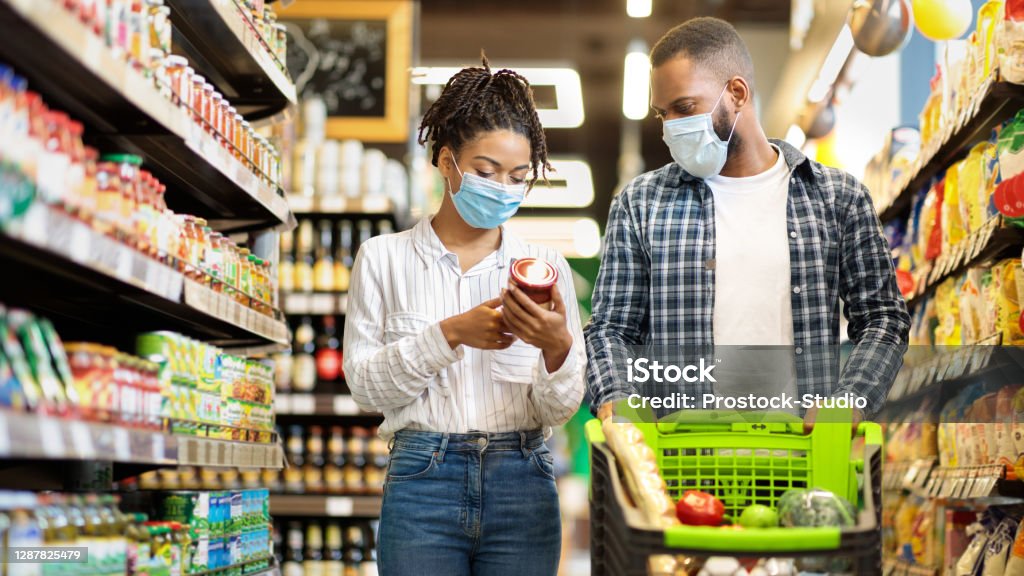 African Family In Shop Buying Groceries Wearing Face Masks, Panorama African Family Couple In Shop Buying Groceries Wearing Face Mask Choosing Food Goods Walking With Shopping Cart In Supermarket Store. Customers Buy Essentials During Pandemic. Panorama, Free Space Supermarket Stock Photo