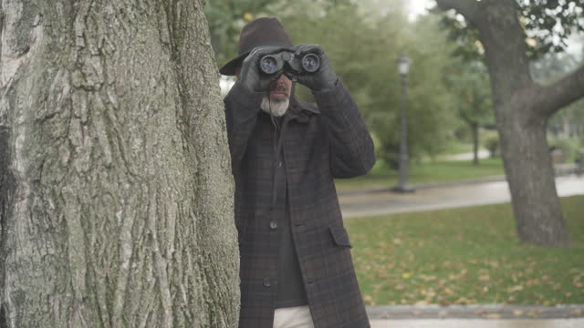 Professional intelligencer or spy standing behind tree and using binoculars. Portrait of confident Caucasian undercover agent or detective hiding in park for spying after target.