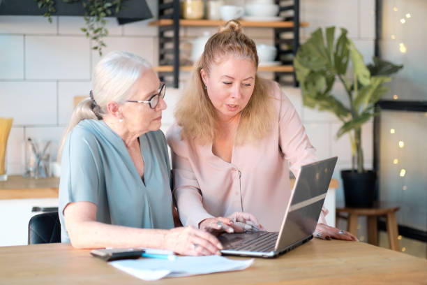 daughter helps her elderly mother figure it out online with her personal account. Woman teaching senior mother to use internet at home. Senior woman with her daughter looking at modern gadget indoors stock photo