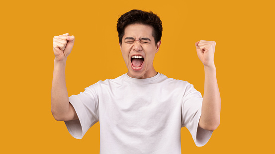 Winner Concept. Studio portrait of overjoyed asian man yelling with clenched fists raised up in the air, isolated over orange background. Excited guy cheering and screaming, celebrating victory