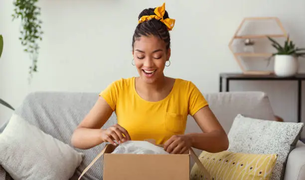 Quick Delivery Service Concept. Happy black lady received package, unpacking cardboard box, sitting on the sofa in living room at home, copy space. Female buyer satisfied with online shopping purchase