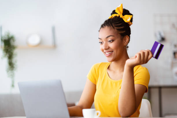 Black woman using computer and credit card at home Easy Payment And Cashback Concept. Portrait of excited african american young lady holding credit card in hand and using laptop, sitting at table at home, typing on keyboard, blurred background cyber monday stock pictures, royalty-free photos & images