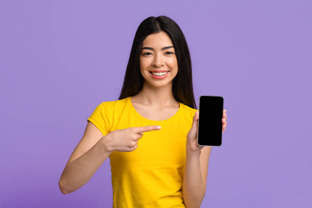 istock Check This. Cheerful Asian Lady Pointing At Smartphone With Black Blank Screen 1287832136