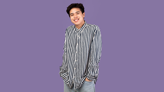 Male Portrait. Smiling asian guy standing and posing at studio, holding hands in pockets, looking at camera, wearing striped shirt, posing isolated over purple studio background. Casual teenage model