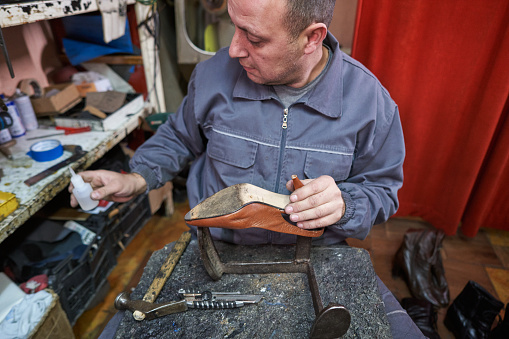Mid adult cobbler hand repairing shoeshigh heels in his messy workshop, while wearing protective workwear