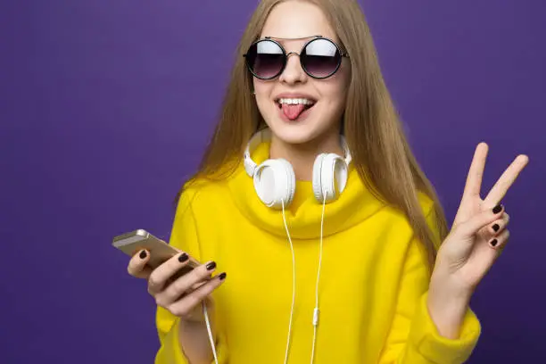 Hello  peace. Young beautiful girl in yellow sweatshirt and glasses. Holds the phone in his hand and headphones around his neck