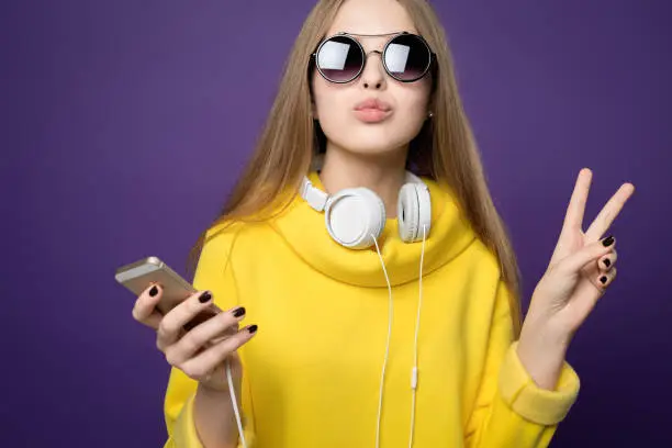 kissing peace. Young beautiful girl in yellow sweatshirt and glasses. Holds the phone in his hand and headphones around his neck