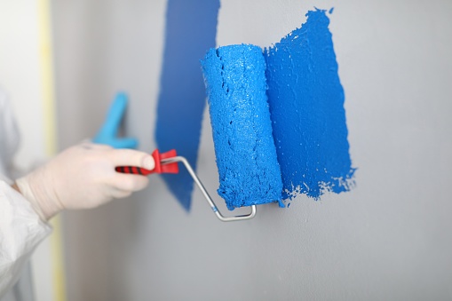 Craftsman holds roller and paints white wall blue. Painter services concept
