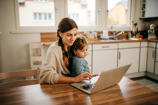 Young mother with headset and a laptop is working from her home office. Her son is sitting in her lap and observing what is she doing. Horizontal photo.