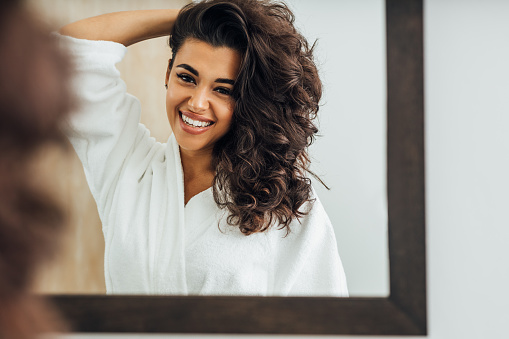 Beautiful smiling woman wearing a bathrobe and holding her hair while standing in front of a mirror