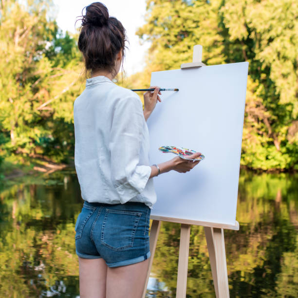 woman artist in summer in a park by lake, background forest water shore trees, draws a picture, view from back, in her hand palette with paints, a brush and an easel. Jeans clothes white shirt. stock photo