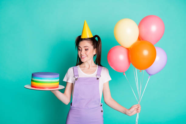 Photo of charming girl hold balloons look cake wear violet overall paper cone headwear isolated turquoise color background Photo of charming girl hold balloons look cake wear violet overall paper cone headwear isolated turquoise color background. woman birthday cake stock pictures, royalty-free photos & images