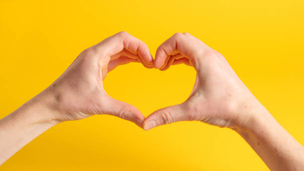 istock Two hand forming a heart shape with the fingers. Heart shaped hands on yellow background. Hands making a heart 1287880937