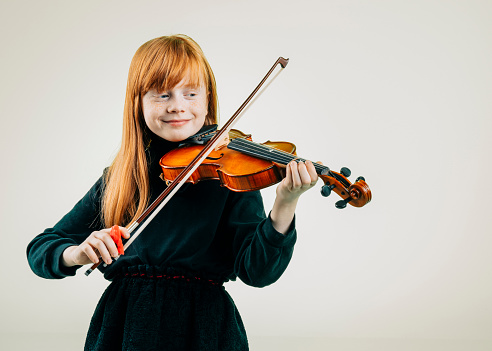 Beautiful young girl with red hair standing confidently enjoying playing the violin. The young musician is smiling proudly while practicing on the instrument.