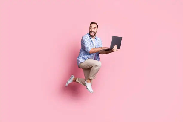 Full length photo of excited guy run jump hold computer wear plaid shirt pants sneakers isolated pink color background.