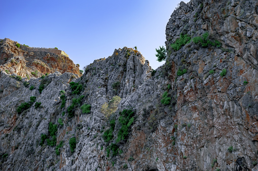 Rock with ancient walls of Alanya Castle (Turkey) on top - bottom view. A steep cliff with green plants on a stone slope against a blue sky