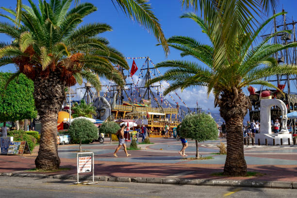 Turkey, Alanya - October 22, 2020: Tropical exotic promenade in Alanya Marina (Alanya Yat Limani). Palm trees grow on the city street against the background of vintage antique pirate ships