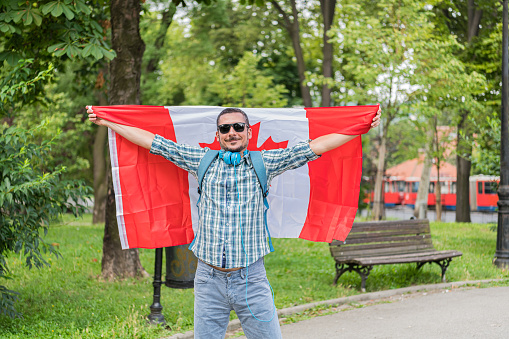 Handsome Tourist With Sunglasses is Enjoying in Summer Day While Walking Through the Public Park and Enjoying in Music Using Headphones, Holding Canadian Flag.