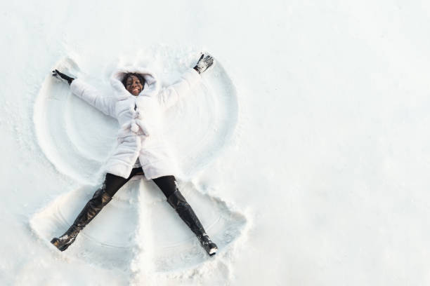 black girl in fur coat and high top boots lies on white glade and draws snow angel picture with arms and legs top view