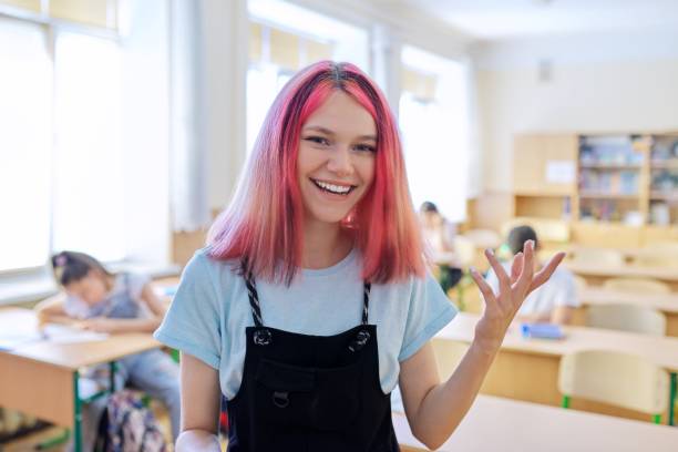 Talking gesturing smiling teenage student at lesson in classroom, trendy beautiful girl looking at camera, background students sitting at desks