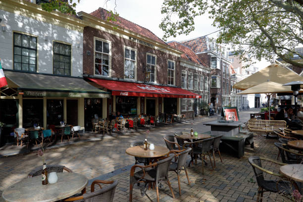 Delft, The Netherlands - October 10, 2019: On the ground floor of the houses there are cafes and restaurants, outside there are also visible tables and chairs of these restaurants. Delft is a popular tourist destination. Many tourists come here and find in this city a summary of the architecture, art and landscape of the Netherlands. Many historical buildings have been preserved in the old part of the city. There are many narrow streets and many of them are canals, the banks of which are connected by small bridges. The city is known for its connections to art, culture and science.