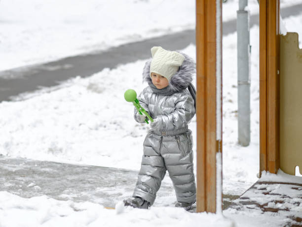 uma garotinha alegre em um macacão prateado quente no playground. o bebê está segurando um brinquedo de bola de neve. - playground snow winter little girls - fotografias e filmes do acervo