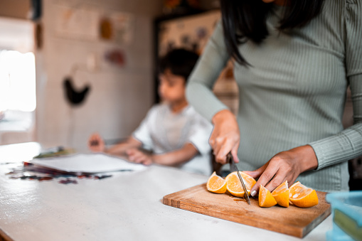 Mother and son are in the kitchen. She is slicing orange, the focus is on her. Son is in the background writing homework
