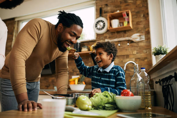 Here dad, try this food which I made! Happy African American boy having fun while feeding his father in the kitchen. family dinners and cooking stock pictures, royalty-free photos & images