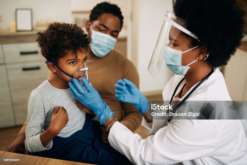 Small black boy receiving asthma treatment while doctor is vising him at home due to COVID-19 pandemic. African American pediatrician using nebulizer during inhaling therapy of a small boy due to coronavirus pandemic. Asthmatic Stock Photo