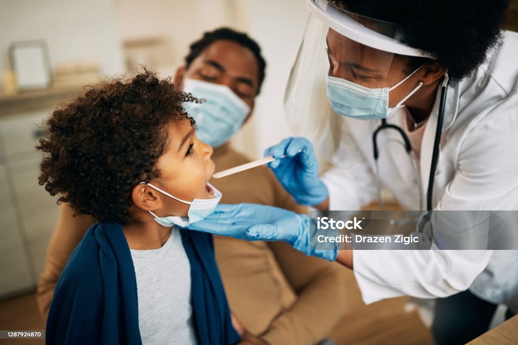 African American doctor with face mask examining boy's throat during a home visit. Family doctor examining throat of a small black boy while visiting him at home during coronavirus pandemic. Doctor Stock Photo