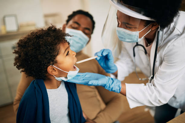African American doctor with face mask examining boy's throat during a home visit. Family doctor examining throat of a small black boy while visiting him at home during coronavirus pandemic. pandemic illness stock pictures, royalty-free photos & images