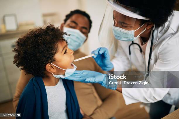 African American Doctor With Face Mask Examining Boys Throat During A Home Visit Stock Photo - Download Image Now