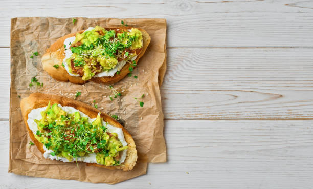 Two avocado sandwiches, tomato, micro greens and sesame seeds on baguette toast with cream cheese on a paper lining, white wooden table. Top view with copy space stock photo