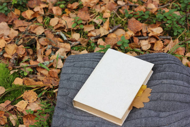 book with a bookmark of leaves on a knitted wool blanket on the grass with autumn leaves.