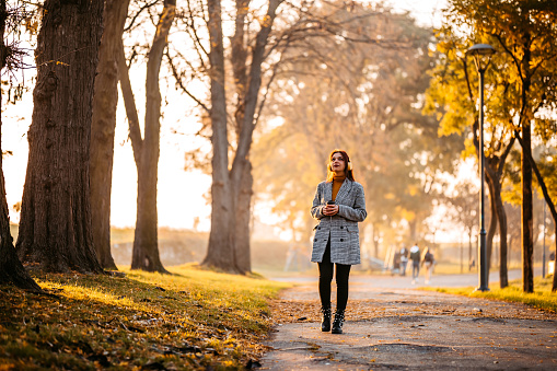 Beautiful young woman drinking coffee and listening music while walking in public park at sunny day.