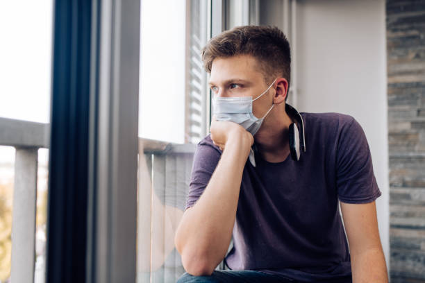 istock Young man with a face mask sitting in a home quarantine and looking through the window 1287950064