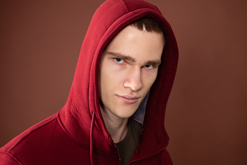 Close up studio portrait of 18 year old man with short brown hair in maroon hooded shirt on brown background