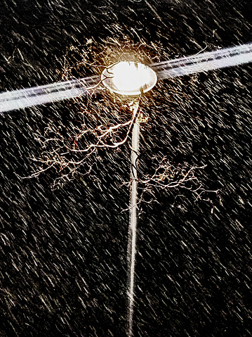 The light of the lantern illuminates a Blizzard, a snow storm. The street lamp shines through a strong blizzard. Wintery nightly, town-like painting. Under the bright light of a lot of snowflakes fall