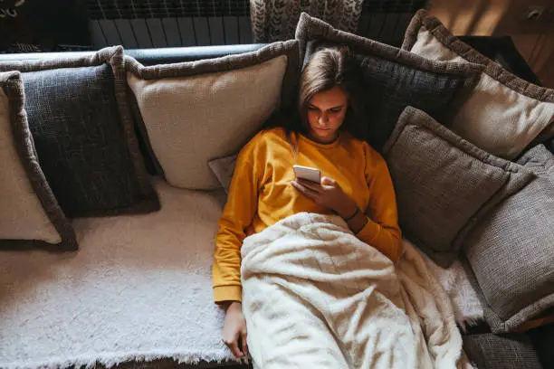 Photo of Young woman lying on sofa and using cell phone at home