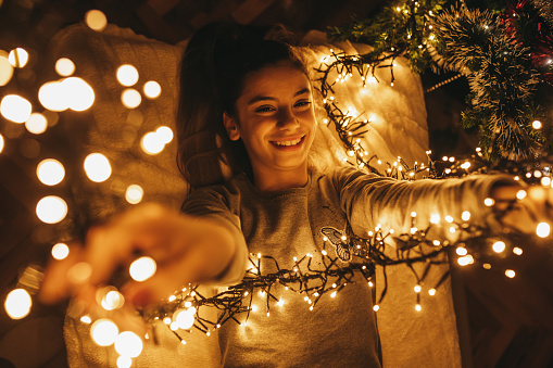 Beautiful young woman decorating Christmas tree and playing with Christmas lights at home